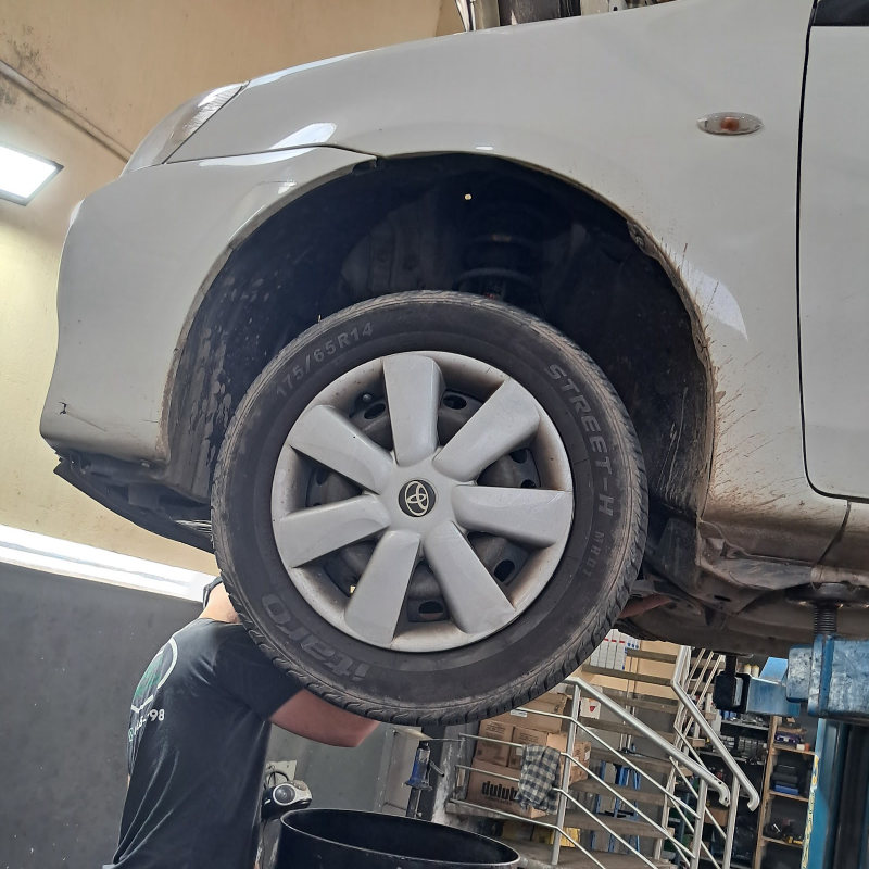 Mecânico embaixo de um carro prata erguido no elevador, com o tambor de óleo ao lado